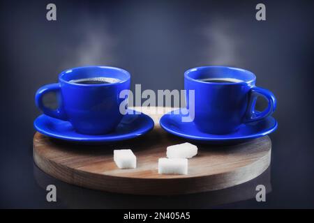 Zwei Tassen frischen Kaffees auf einem Holzständer. Dampf aus einer Kaffeetasse. Blaue Tassen mit einem heißen Getränk auf dunklem Hintergrund neben dem Zucker. Speicherplatz kopieren Stockfoto