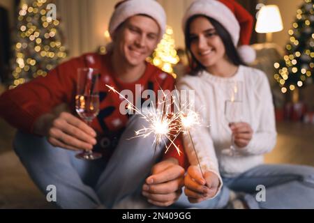 Ein Paar in Weihnachtsmannhüten mit Glitzer und Champagnergläsern, konzentriert euch auf das Feuerwerk. Weihnachtsfeier Stockfoto