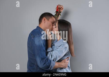 Ein glückliches Paar küsst sich unter Mistelzweigen auf grauem Hintergrund Stockfoto