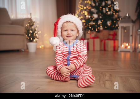 Süßes Baby im Weihnachtsschlafanzug und Weihnachtsmannmütze drinnen Stockfoto