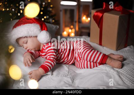 Baby im Weihnachtsschlafanzug und Weihnachtsmannmütze schlafen in der Nähe der Geschenkbox auf dem Bett drinnen Stockfoto