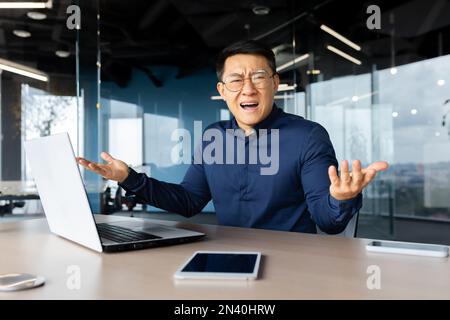 Porträt eines frustrierten und verärgerten Chefs im Büro, eines asiatischen Mannes, der in die Kamera schaut und verärgert schreit, eines Mannes am Arbeitsplatz, der ein Notebook verwendet und mit dem Arbeitsergebnis unzufrieden ist. Stockfoto