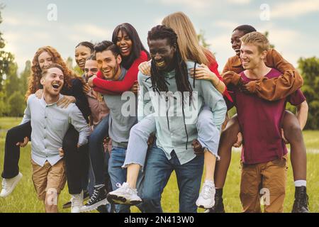 Eine große Gruppe von Freunden aus verschiedenen Rassen, die das Wochenende im Park genießen und gemeinsam Spaß beim Huckepack haben - Vertrauen, Glück und Freundschaft Lebensstil Contra Stockfoto