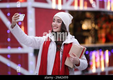 Glückliche junge Frau mit Geschenkbox, die abends Selfie auf der Wintermesse macht. Weihnachtsfeier Stockfoto