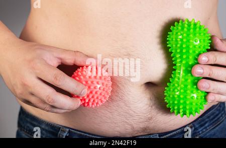 Ein Mann hält zwei Anti-Cellulite-Massagegeräte vor dem Hintergrund eines dicken Bauches. Fettverbrennung mit Verfahren. Schneller Gewichtsverlust. Stockfoto