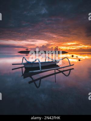 Ein vertikales Bild eines Boots auf dem Wasser bei Sonnenuntergang in Pantai Karang, Sanur, Bali, Indonesien Stockfoto