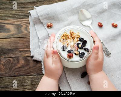 Natürlicher, hausgemachter Joghurt in einem Glasgefäß. Gesunde Nahrung zum Frühstück in Kinderhänden. Ein Kind hielt einen Becher mit Müsli auf Leinentischdecken auf einem Holztisch Stockfoto