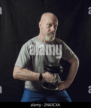 Ein älterer, rasierter Mann mit grauem Bart trainiert seine Arme mit Kurzhanteln. Stockfoto