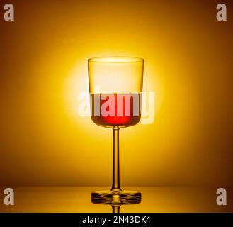 Ein Glas Rotwein auf leuchtendem orangefarbenem Hintergrund spiegelt sich auf einem Spiegeltisch wider. Stockfoto