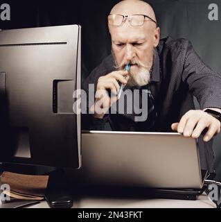 Bei der Arbeit im Büro ist ein ernster älterer Mann mit einem Bart und einem Bleistift besorgt über den Laptop-Bildschirm. Stockfoto