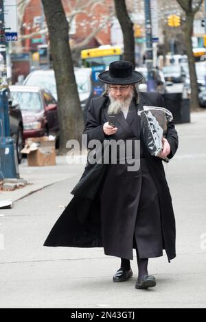 Ein älterer orthodoxer Jude, der aus der Synagoge zurückkehrt, liest sein Klapphandy. In Brooklyn, New York. Stockfoto