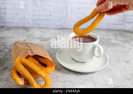 Churros in einer Papiertüte mit einer Tasse heißer Schokolade, typisches spanisches Frühstück, auf steingrauem Hintergrund Stockfoto