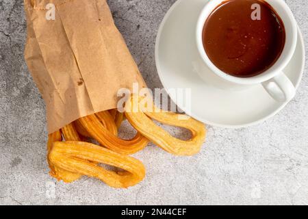 Churros in einer Papiertüte mit einer Tasse heißer Schokolade, typisches spanisches Frühstück, auf steingrauem Hintergrund Stockfoto