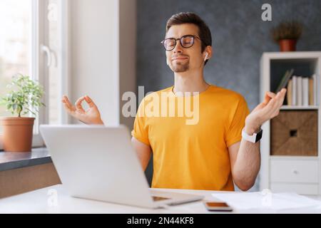 Ein junger, friedlicher und aufmerksamer Mann bleibt ruhig, lächelt, entspannt, meditiert, während er von zu Hause aus mit einem Laptop arbeitet und Yoga-Atemübungen durchführt. Online-Bildung, soziale Distanzierung. Bleib zu Hause, Covid Stockfoto