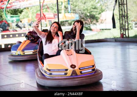 Zwei junge Frauen auf einer lustigen Autofahrt im Vergnügungspark, die ein Selfie mit dem Smartphone machen. Stockfoto
