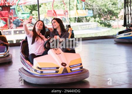 Zwei junge Frauen auf einer lustigen Autofahrt im Vergnügungspark, die ein Selfie mit dem Smartphone machen. Stockfoto