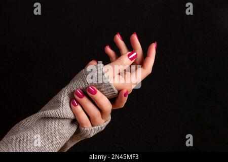 Weibliche Hände in grauem Strickpullover mit schöner Maniküre – Viva Magenta, pinkfarbene Nägel auf dunkelschwarzem Hintergrund. Nagelpflegekonzept Stockfoto