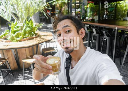 Junger venezolanischer Latino-Mann mit Dreadlocks, der in einem Restaurant steht, ein Selfie-Porträt mit einem Kaffee in der Hand macht, in die Kamera schaut, perspektiv Stockfoto