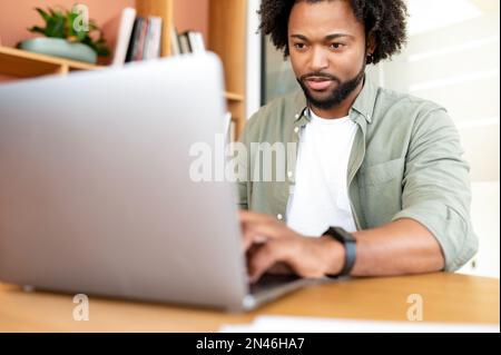 Der gutaussehende junge Geschäftsmann benutzt in Innenräumen ein Notebook. Ein afroamerikanischer Büroangestellter tippt auf der Tastatur, antwortet auf E-Mails, männlicher Freiberufler in Freizeitkleidung, der Remote arbeitet Stockfoto