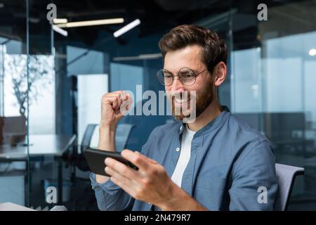 Ein Mann im Büro, der Videospiele auf einem Smartphone spielt, ein Geschäftsmann, der mit dem Gewinnen zufrieden ist, freut sich und hält die Hand in Triumph-Geste, der Boss im Hemd ist mit Erfolg und Ergebnis zufrieden. Stockfoto