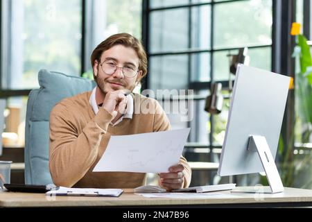 Porträt eines jungen Designers, Architekten, Ingenieurs, der in einem Büro sitzt und an einem Computer arbeitet. Er hält das Kinn mit der Hand, hält Dokumente, einen Plan, ein Projekt in seinen Händen. Er lächelt in die Kamera. Stockfoto