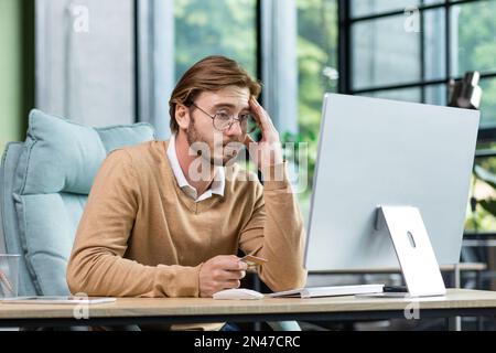 Kreditkartentransaktion fehlgeschlagen. Ein verärgerter junger Mann sitzt im Büro am Tisch, schaut auf den Computer, hält eine Kreditkarte in der Hand. Enttäuscht lehnte er sich auf seinen Arm. Stockfoto