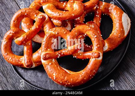 Nahaufnahme von weichen Brezeln in Form von Knoten gebacken und mit Salz auf schwarzem Teller auf dunklem Holztisch bestreut, horizontale Ansicht von oben Stockfoto