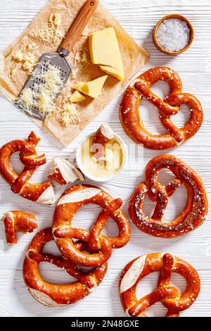 Weiche Brezeln in Form von Knoten gebacken und mit Salz auf weißem Holztisch mit Käsesauce und geriebenem Käse bestreut, vertikale Ansicht von oben Stockfoto