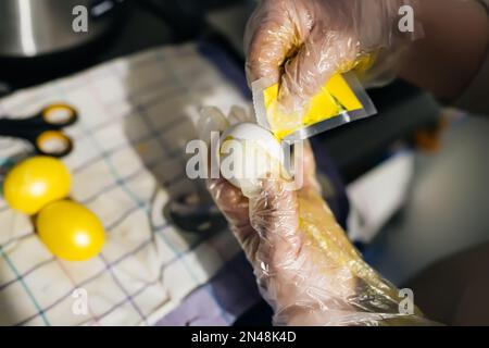 Ostereier mit gelben Farbstoffen lackieren. Die Hände in Polyethylen-Handschuhen halten ein Hühnerei, das mit Farbe aus dem Beutel bedeckt ist. Teil des Stockfoto