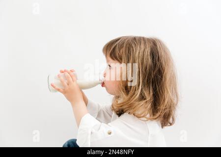 Ein hübsches Mädchen mit lockigem Haar und blauen Augen trinkt Milch aus einer Flasche. Weißer Hintergrund. Stockfoto