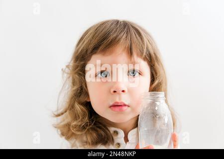 Ein hübsches Mädchen mit lockigem Haar und blauen Augen trinkt Milch aus einer Flasche. Weißer Hintergrund. Stockfoto