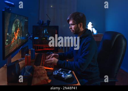 Konzentrierter Pro-Gamer Spielt Spiel, Nimmt Am Online-Turnier Teil, Sitzt Am Computer Zu Hause. Schwaches Licht, Neonfarbe, Seitenansicht. Professionelle Cybersportler trainieren Online-Spiel auf seinem PC Stockfoto