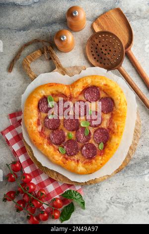 Herzförmige Pizza am Valentinstag mit Mozzarella, Peperoni und Basilikum, Weinflasche, zwei Weingläsern, Geschenkbox auf hellgrauem Hintergrund. Eine Idee für Romantiker Stockfoto