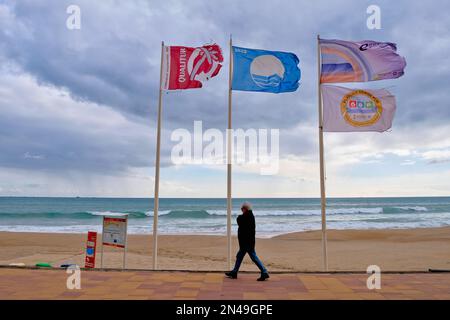 Villajoyosa Costa Blanca Spanien 02.08.2023 Blue Flag Beach. Mann, der im Winter an der gefliesten Strandpromenade spaziert. Drei Pole mit Bannern flowin Stockfoto