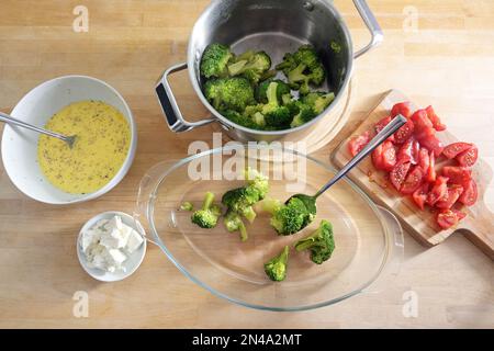 Brokkoli in einem Topf, geschnittene Tomaten und Zwiebeln, Fetakäse und eine Eiermischung werden in ein Glasbackgericht gefüllt, Zubereitung für ein Gemüse Stockfoto