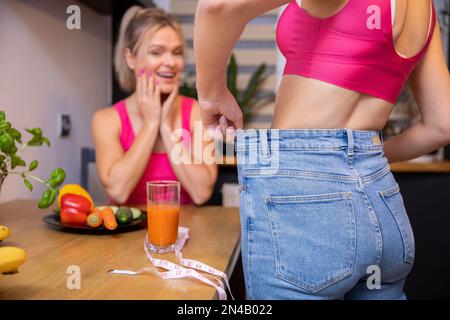 Eine Frau bewundert die Gewichtsabnahme ihrer Freundin, während sie in der Küche ist Stockfoto