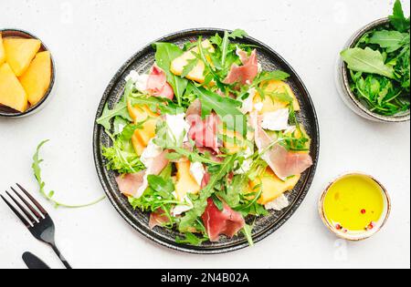 Köstlicher Melonensalat mit Cantaloupe, Prosciutto, Weichkäse und Rucola auf weißem Tischhintergrund, Draufsicht Stockfoto