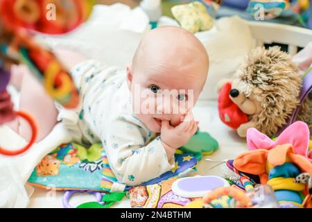 Ein süßes, kleines, neugeborenes Mädchen liegt auf einem Bett inmitten bunter Spielzeuge für Babys. Das erste pädagogische Spielzeug für die sensorische Entwicklung. Stockfoto