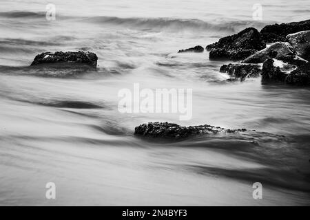 Eine Graustufenaufnahme von Meereswellen, die die Felsen am Strand stürzen. Stockfoto