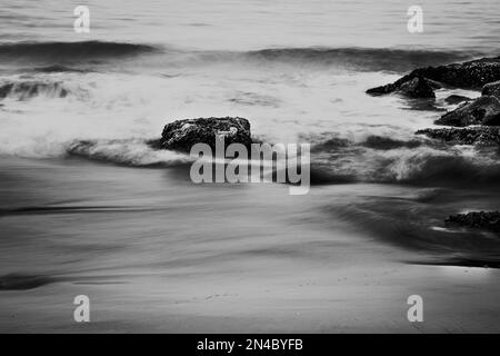 Eine Graustufenaufnahme von Meereswellen, die die Felsen am Strand stürzen. Stockfoto