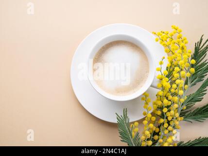 Ein Frühlingsbouquet mit gelben Mimosablüten und eine Tasse Kaffee-Cappuccino auf beigem Hintergrund. Konzept vom 8. März, fröhlicher Frauentag. Draufsicht und Stockfoto