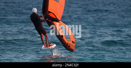 Ein Mann trägt Flügelfolien mit handgeführten aufblasbaren Flügeln und Tragflächensurfbrettern in einem blauen Ozean, reitet auf einem Windflügelbrett, surft auf den Wellen Stockfoto