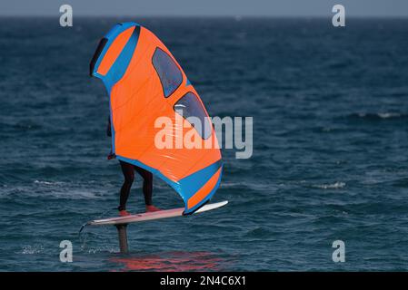 Ein Mann trägt Flügelfolien mit handgeführten aufblasbaren Flügeln und Tragflächensurfbrettern in einem blauen Ozean, reitet auf einem Windflügelbrett, surft auf den Wellen Stockfoto