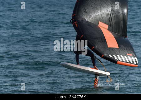 Ein Mann trägt Flügelfolien mit handgeführten aufblasbaren Flügeln und Tragflächensurfbrettern in einem blauen Ozean, reitet auf einem Windflügelbrett, surft auf den Wellen Stockfoto