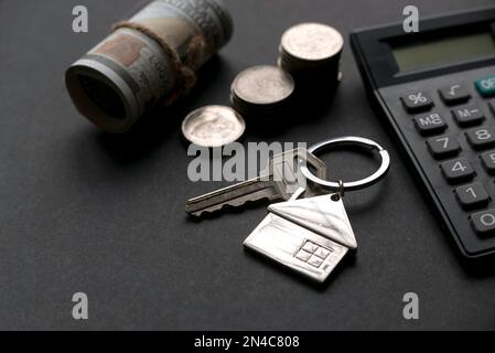 Hausschlüssel mit Taschenrechner, Geld und einem Stapel Münzen auf schwarzem Hintergrund. Stockfoto