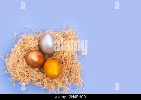 Mehrfarbige bemalte Eier in einem hausgemachten Nest auf blauem Hintergrund, Kopierraum. Ostern minimales kreatives Layout. Frohe Osterferien. Eine Grußkarte. Stockfoto