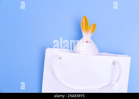 Ostern einkaufen. Weiße Einkaufstasche aus Papier mit weißem Häschen auf blauem Hintergrund. Konzept Ostergeschenke, Vorbereitung, Weihnachtsüberraschung. Stockfoto