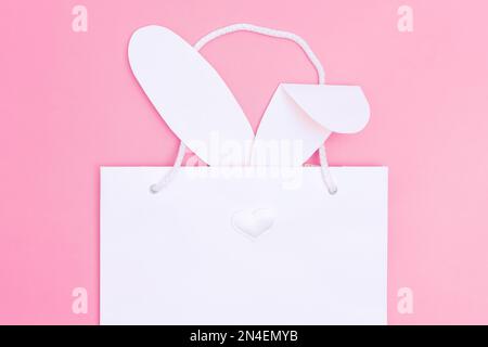 Ostern einkaufen. Weiße Einkaufstasche aus Papier mit weißen Hasenohren auf pinkfarbenem Hintergrund. Konzept Ostergeschenke, Vorbereitung, Weihnachtsüberraschung. Stockfoto