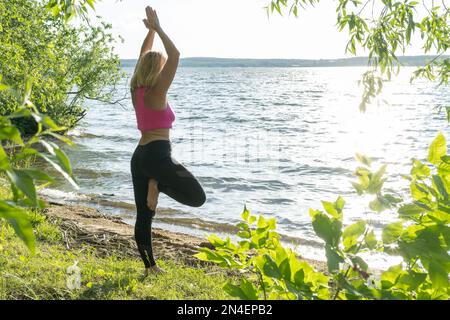 Eine ältere Frau in Sportkleidung, im Yoga stehend, Pose Vrikshasana, Baum Pose auf einer Matte am Seeufer, Blick von hinten. Meditation. Yoga Stockfoto