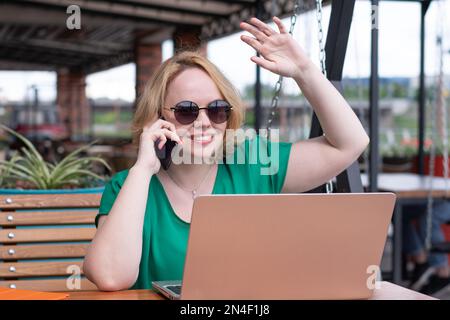 Glückliches Mädchen, das mit Freunden telefoniert, auf dem Laptop surft und mit der Hand Freunde in einem Straßencafé begrüßt. Schülerin in Sonnenbrille, arbeitet an der Stockfoto